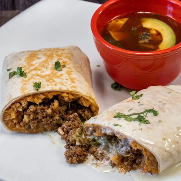 Two burritos, one cut open to show ground beef and rice, served with a red bowl of soup.