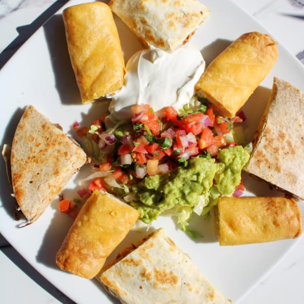 A white plate with quesadilla wedges, taquitos, guacamole, pico de gallo, and sour cream.