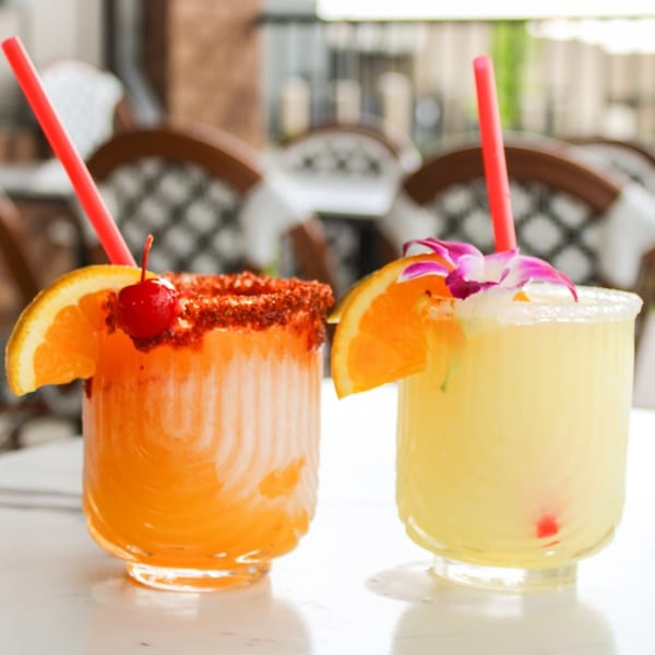 Two vibrant cocktails, one orange with a chili-lime rim and cherry, the other yellow with a sugar rim and orchid.