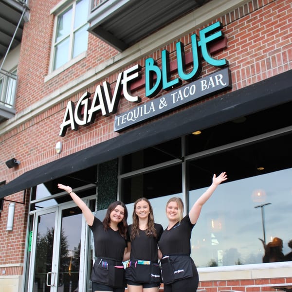 Three smiling women in black uniforms stand outside Agave Blue Tequila & Taco Bar.