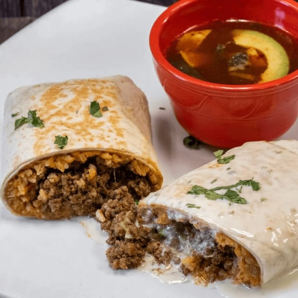 Two burritos, one cut open, revealing ground meat and rice, served with a red bowl of soup.