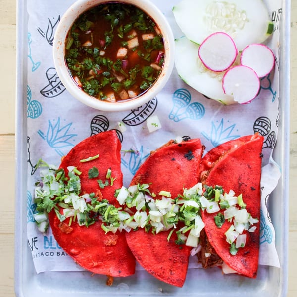 Three red birria tacos topped with diced onions and cilantro, with a side of consommé and sliced radishes.