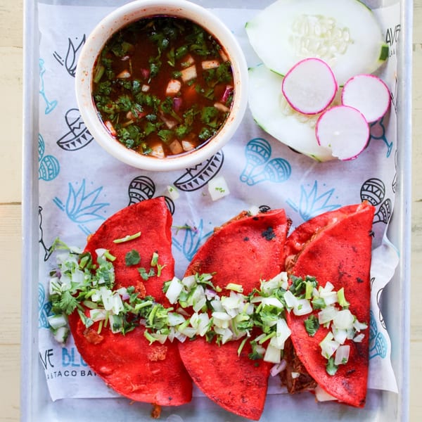 Three red birria tacos with cilantro and onions, a side of consommé, and sliced cucumbers and radishes.