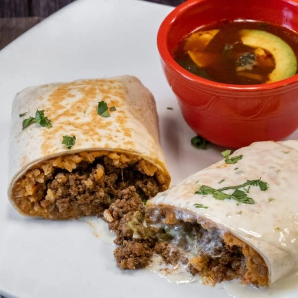 Two burritos, one cut open to show ground beef and rice, with a red bowl of soup.