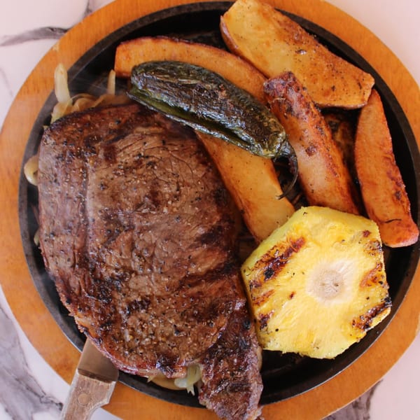 Sizzling steak with grilled pineapple, potato wedges, and a charred jalapeño on a hot plate.