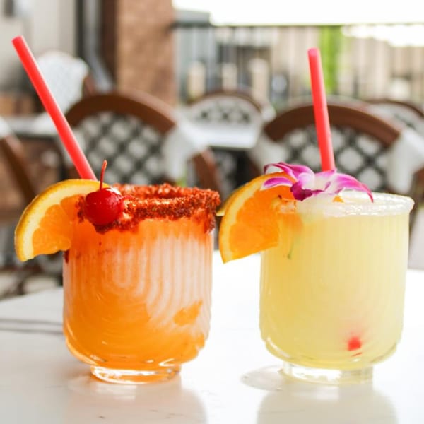 Two vibrant cocktails, one orange with a chili-lime rim and cherry, one yellow with a sugar rim and orchid.