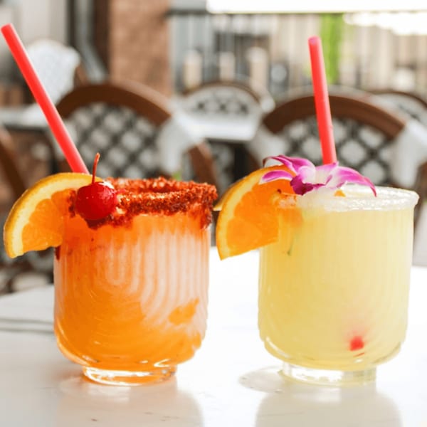 Two vibrant cocktails, one orange with a chili-lime rim and cherry, the other yellow with an orchid.